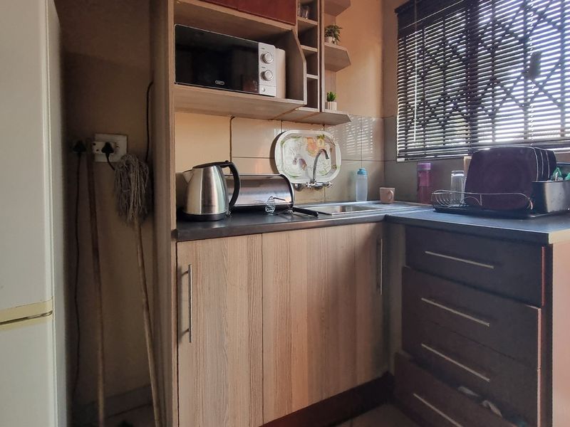 A compact scullery or prep area featuring wood-look cabinetry, laminate countertops, a stainless steel sink, and tiled splashback with window blinds.