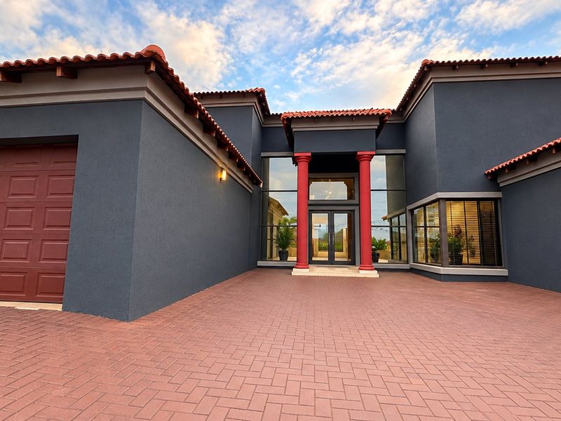 Modern front elevation featuring a paved driveway, double garage, and a striking glass entrance framed by decorative columns and terracotta roof tiles.