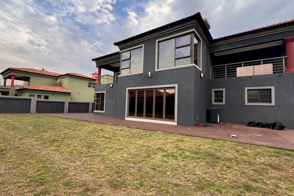 Rear view of a double-story house featuring a paved patio, balcony, and dark grey exterior finish with a spacious lawn.