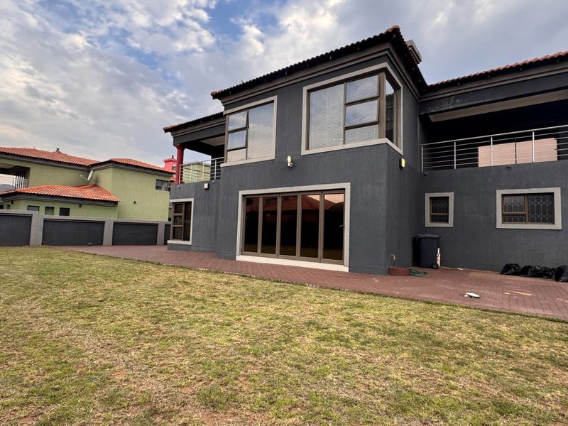 Rear view of a double-story house featuring a paved patio, balcony, and dark grey exterior finish with a spacious lawn.