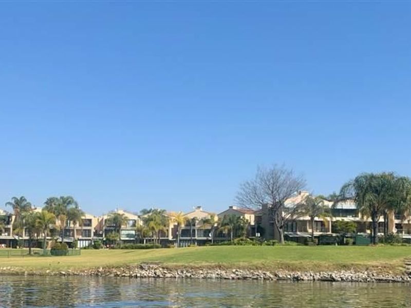 A scenic waterfront view of the townhouse complex, showcasing the well-maintained communal lawns and landscaped garden areas along the water's edge.
