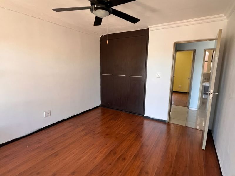 A spacious bedroom featuring dark wood-look laminate flooring, a ceiling fan, and a built-in wardrobe, with an adjoining doorway leading to a bathroom.
