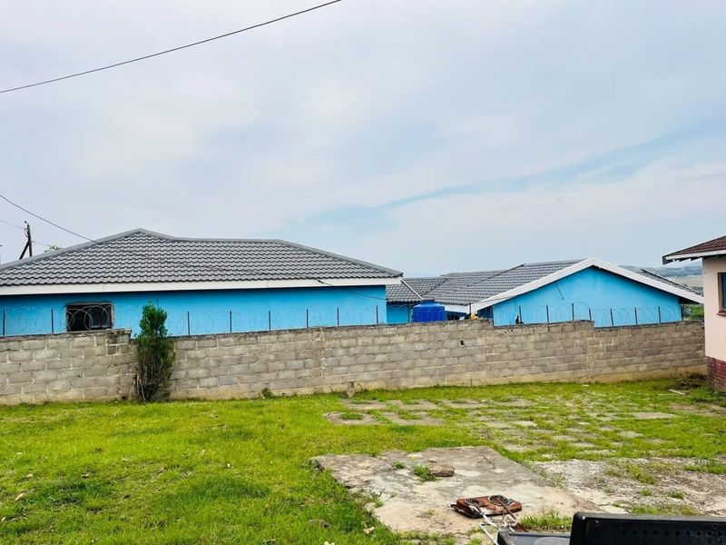 Rear view of the property featuring a blue exterior, tiled roof, concrete block boundary wall with razor wire, and a grassed yard with paved sections.