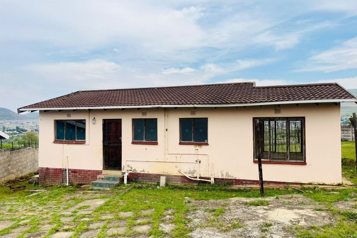 Rear view of a single-story house featuring a tiled roof, security bars, and a paved yard area in need of exterior maintenance and painting.