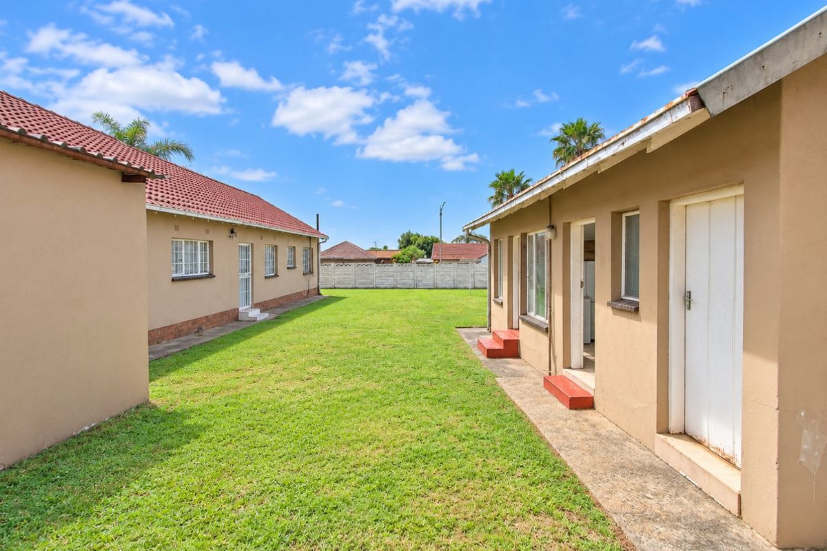 A view of the rear garden area situated between two residential structures, featuring a lawn, concrete pathways, and perimeter boundary walling.