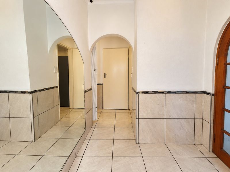 A bright, tiled hallway featuring an arched doorway, wall-mounted mirror, and decorative tile wainscoting, providing a clean and neutral transition space.