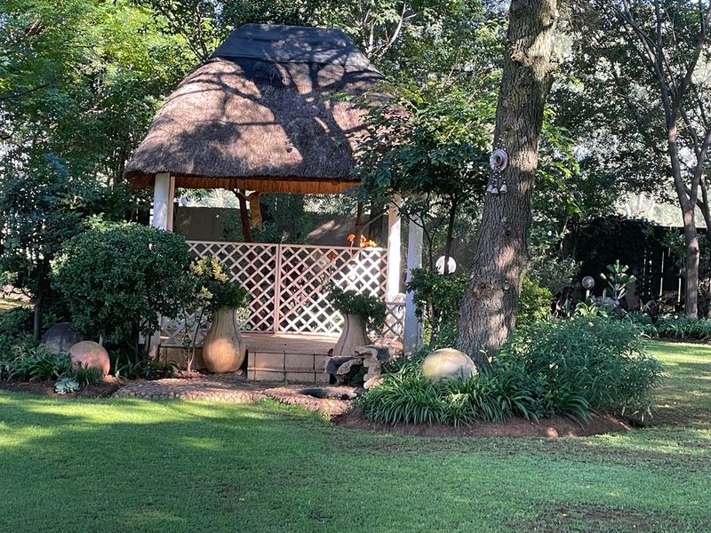 A tranquil garden setting featuring a traditional thatched gazebo with lattice screening, surrounded by mature trees and well-maintained lawn.