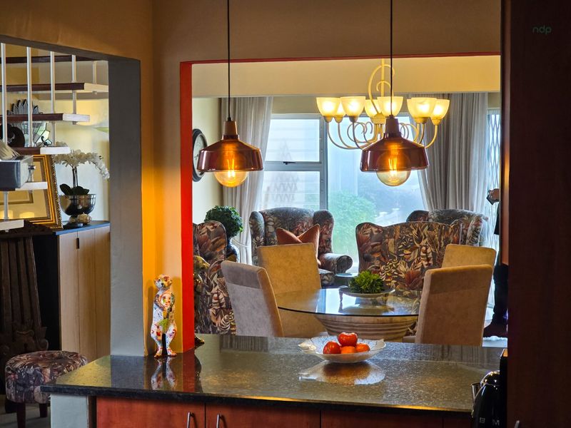 An open-plan living and dining area featuring a granite-topped kitchen counter, elegant pendant lighting, and a classic chandelier, creating a warm and connected living space.