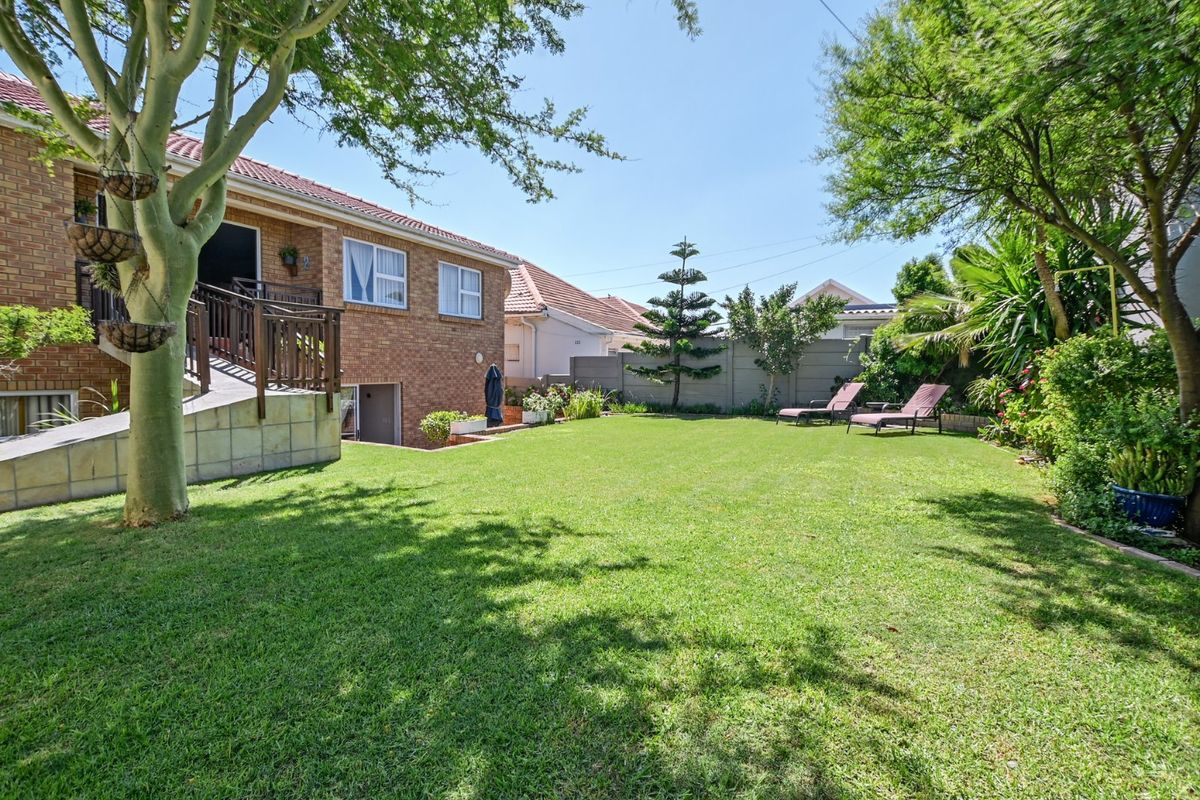 A well-maintained rear garden featuring a spacious lawn, mature trees, and a raised patio area with timber balustrades attached to the brick house.