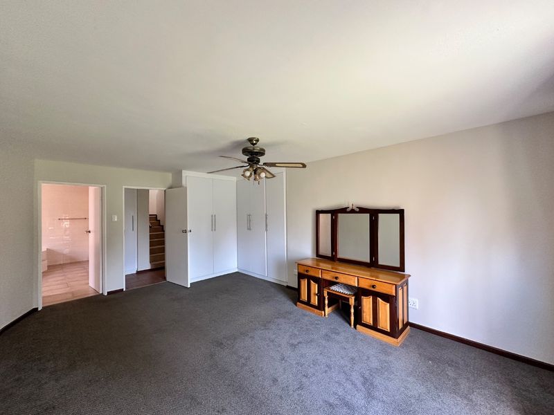 A spacious bedroom featuring built-in cupboards, a ceiling fan, and neutral carpeting, with direct access to an adjoining bathroom and hallway.