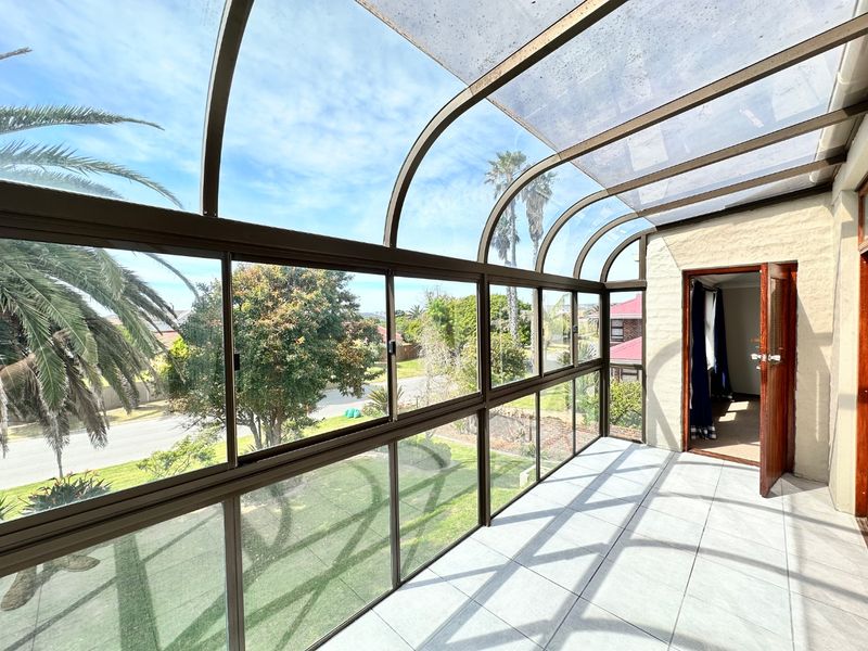 A bright, enclosed patio featuring a curved polycarbonate roof, tiled flooring, and expansive aluminium windows offering views of the surrounding garden and street.
