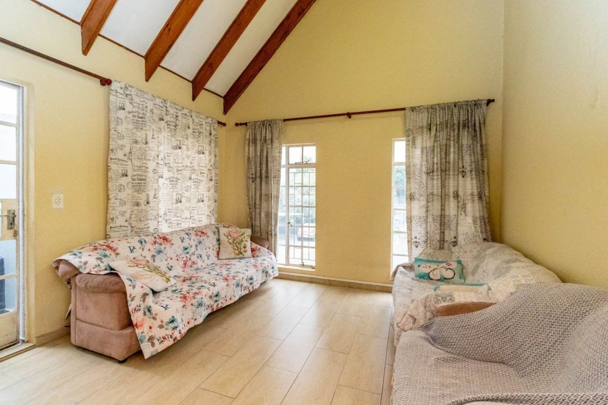 A bright living room featuring a high vaulted ceiling with exposed timber beams, tiled flooring, and large windows providing natural light.