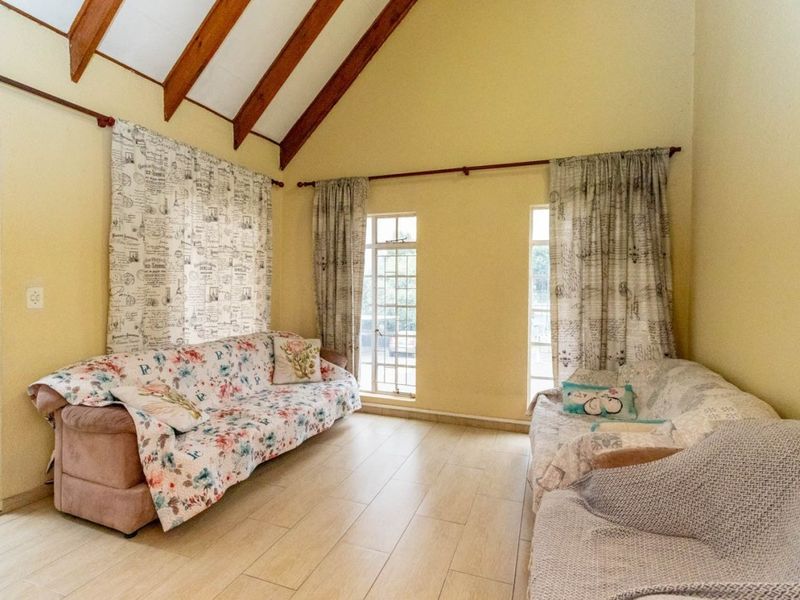 A bright living room featuring a high vaulted ceiling with exposed timber beams, tiled flooring, and large windows providing natural light.