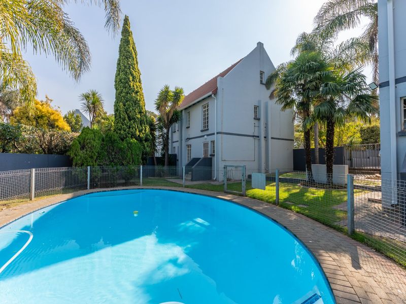 A well-maintained swimming pool area featuring a paved surround, safety fencing, and established garden landscaping set against the townhouse exterior.