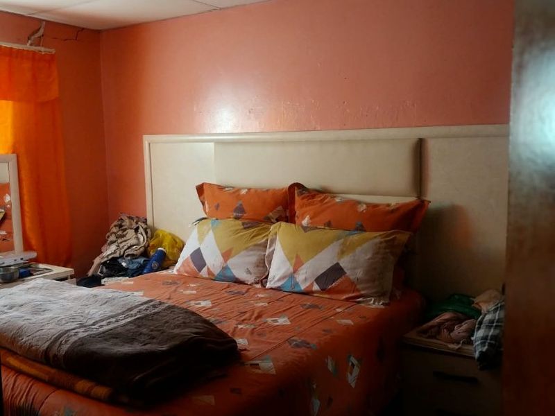 A bedroom featuring a built-in headboard and drop ceiling, showing signs of wear including wall cracking and ceiling panel irregularities.