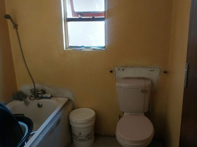 A dated bathroom featuring a bathtub with handheld shower mixer, a toilet, and a small casement window, requiring cosmetic updates and repairs.