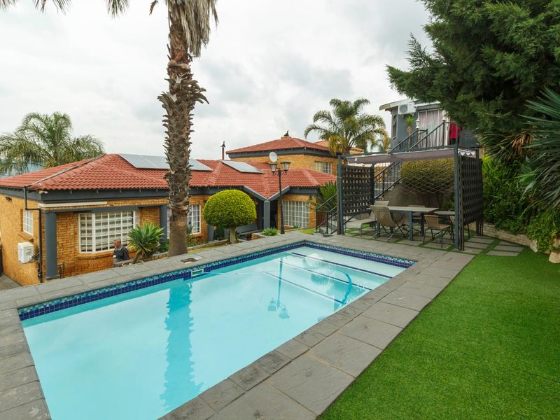 Outdoor pool area featuring a swimming pool with stone coping, artificial lawn, and a covered pergola, set against a brick house with solar panels.