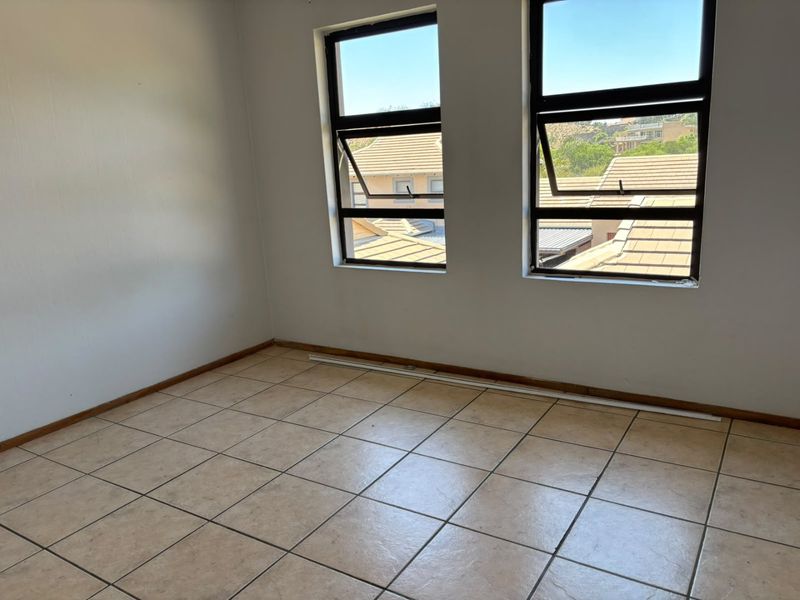 An empty bedroom featuring tiled flooring and dual aluminium-framed windows with views of the surrounding residential area.