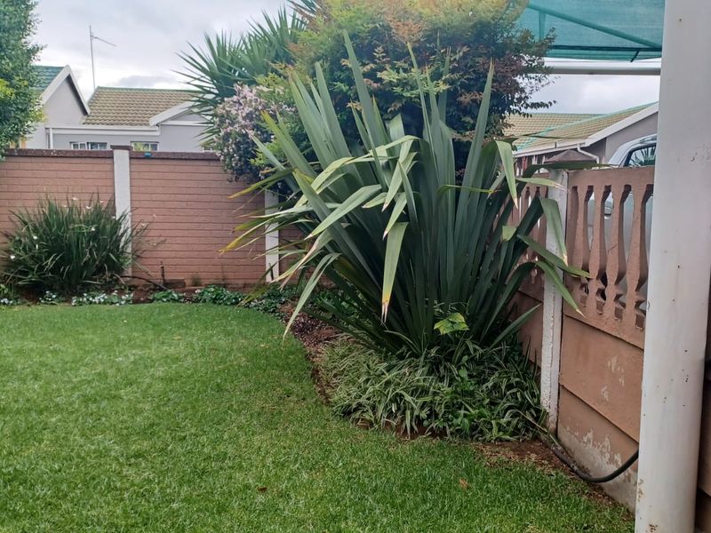 A well-maintained residential garden featuring a lawn, established garden beds, and a combination of brick and precast concrete boundary fencing.