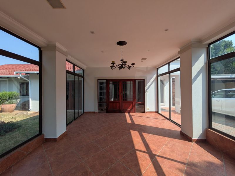 A bright, enclosed patio area featuring large glass windows, tiled flooring, and a central chandelier, offering a versatile space with views of the garden.