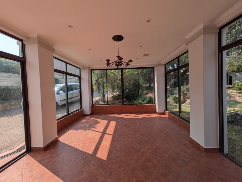 A bright, enclosed patio area featuring large glass windows, tiled flooring, and a decorative chandelier, offering views of the surrounding garden.