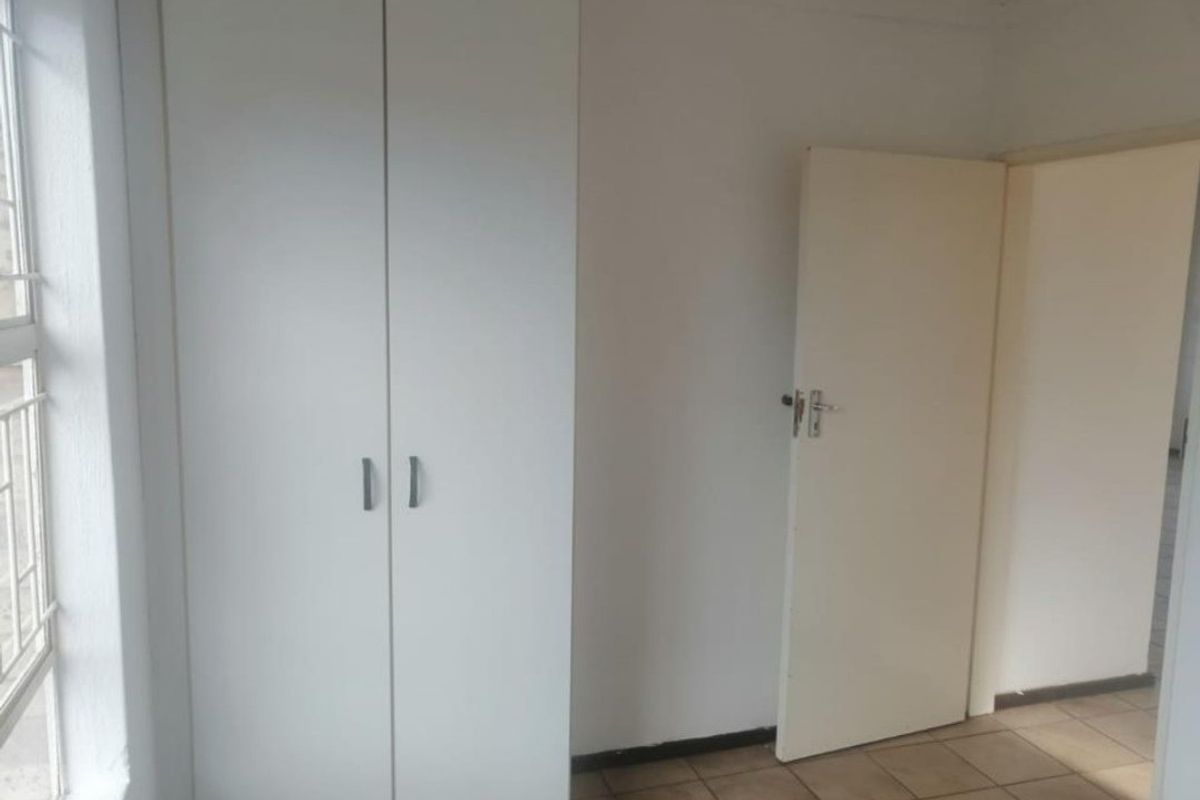 A clean, empty bedroom featuring neutral tiled flooring, a built-in wardrobe, and a window fitted with security bars.