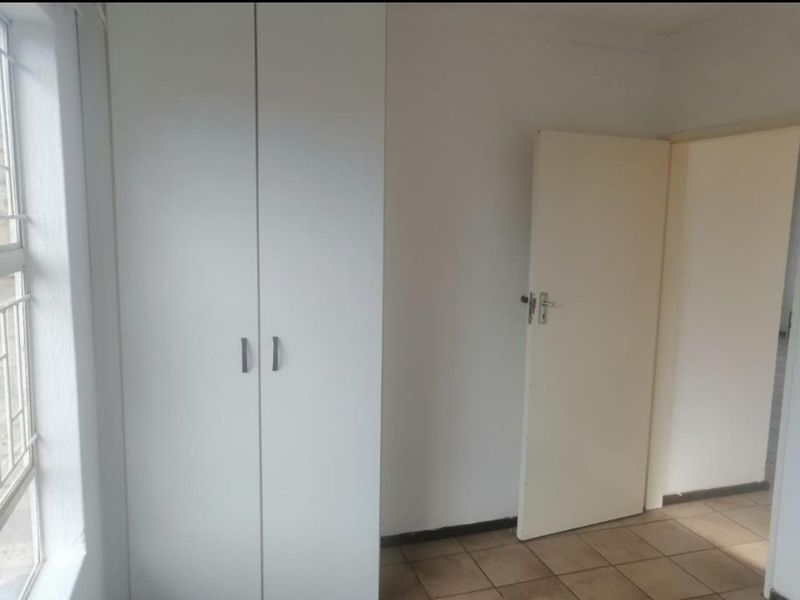 A clean, empty bedroom featuring neutral tiled flooring, a built-in wardrobe, and a window fitted with security bars.