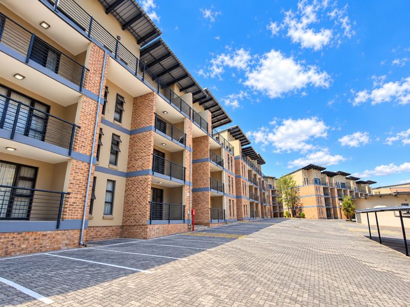 A modern multi-story apartment complex featuring brick facades, private balconies, and a spacious paved parking area under a clear blue sky.