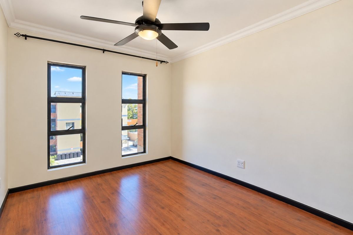 A bright, empty bedroom featuring laminate wood flooring, a ceiling fan, and large windows with views of the surrounding complex.