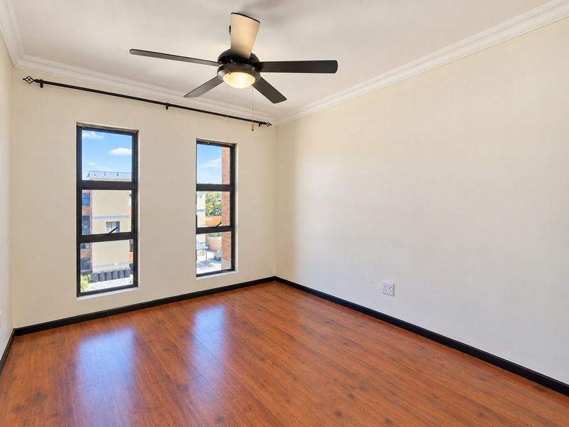 A bright, empty bedroom featuring laminate wood flooring, a ceiling fan, and large windows with views of the surrounding complex.