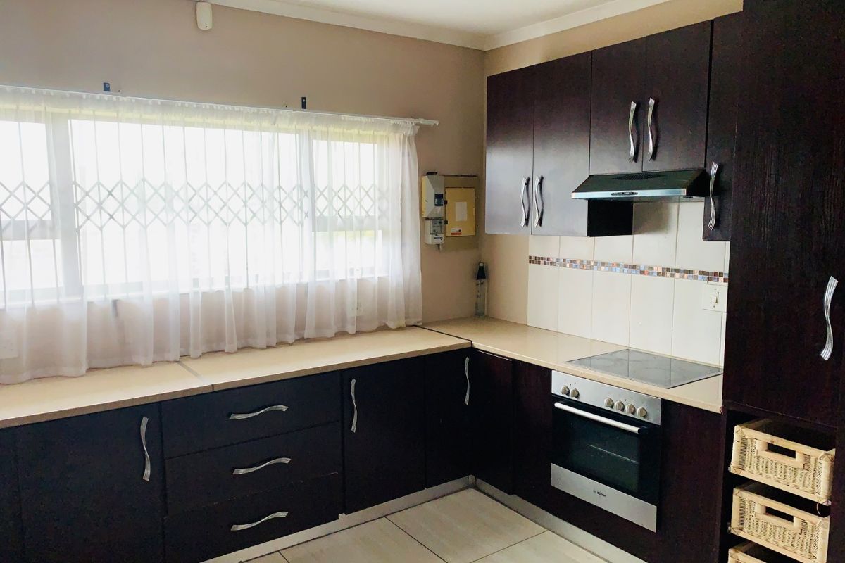 A functional kitchen featuring dark wood cabinetry, built-in oven, electric hob, extractor fan, and tiled flooring with a window security gate.