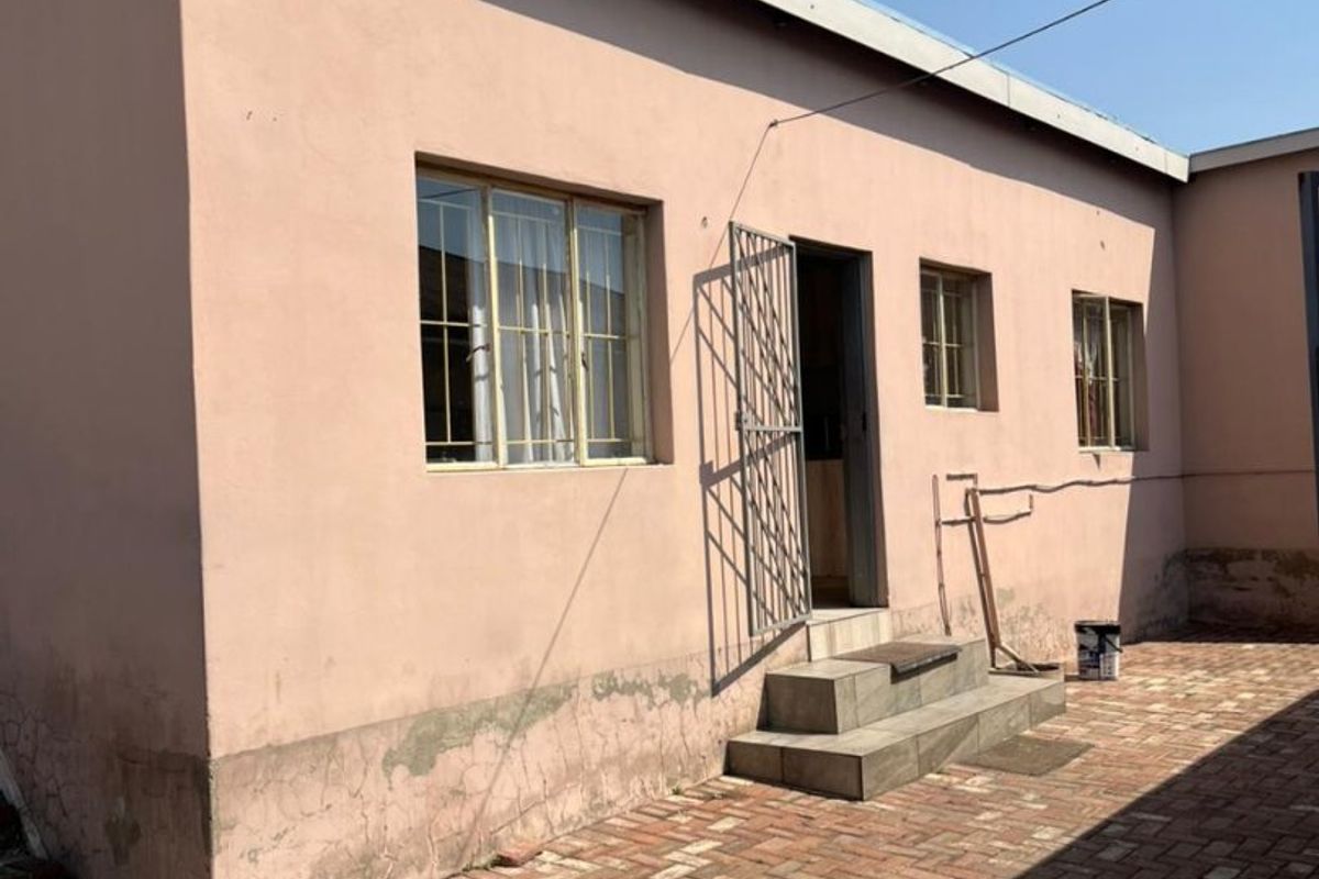 Rear exterior view of the property featuring a paved courtyard, security gate, and tiled entrance steps with visible wear to the exterior wall finish.