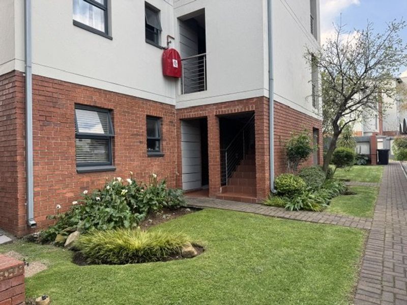 Exterior view of an apartment building featuring a brick and plaster facade, staircase access, and a well-maintained communal garden with paved walkways.