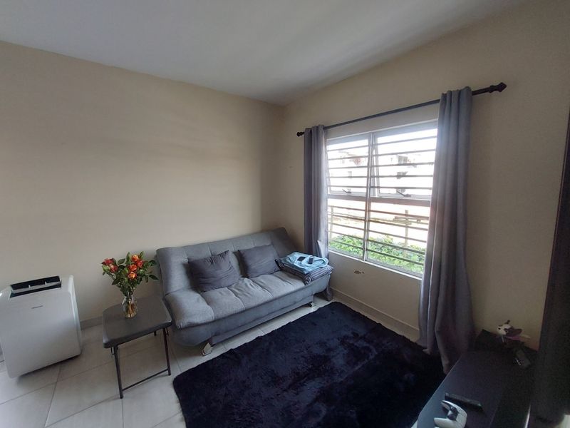 A compact living room featuring tiled flooring and a window fitted with security bars, providing natural light and a functional layout.