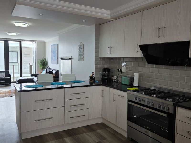 Modern open-plan kitchen and living area featuring light wood cabinetry, granite countertops, gas cooking appliances, and seamless access to an outdoor balcony.