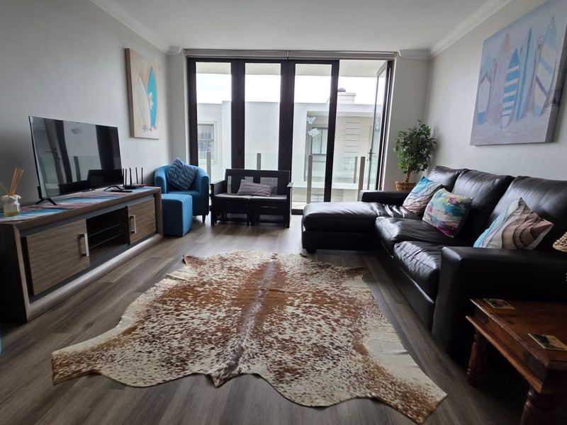 A well-maintained living room featuring modern laminate flooring, coved ceilings, and large sliding glass doors that open onto a balcony.