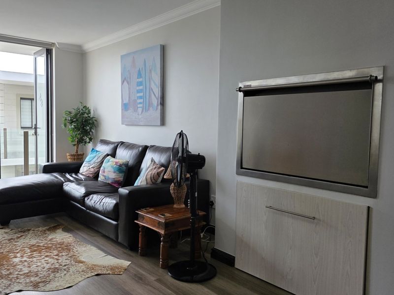 A modern living room featuring laminate flooring, crown molding, and a built-in stainless steel braai, with direct access to a balcony.