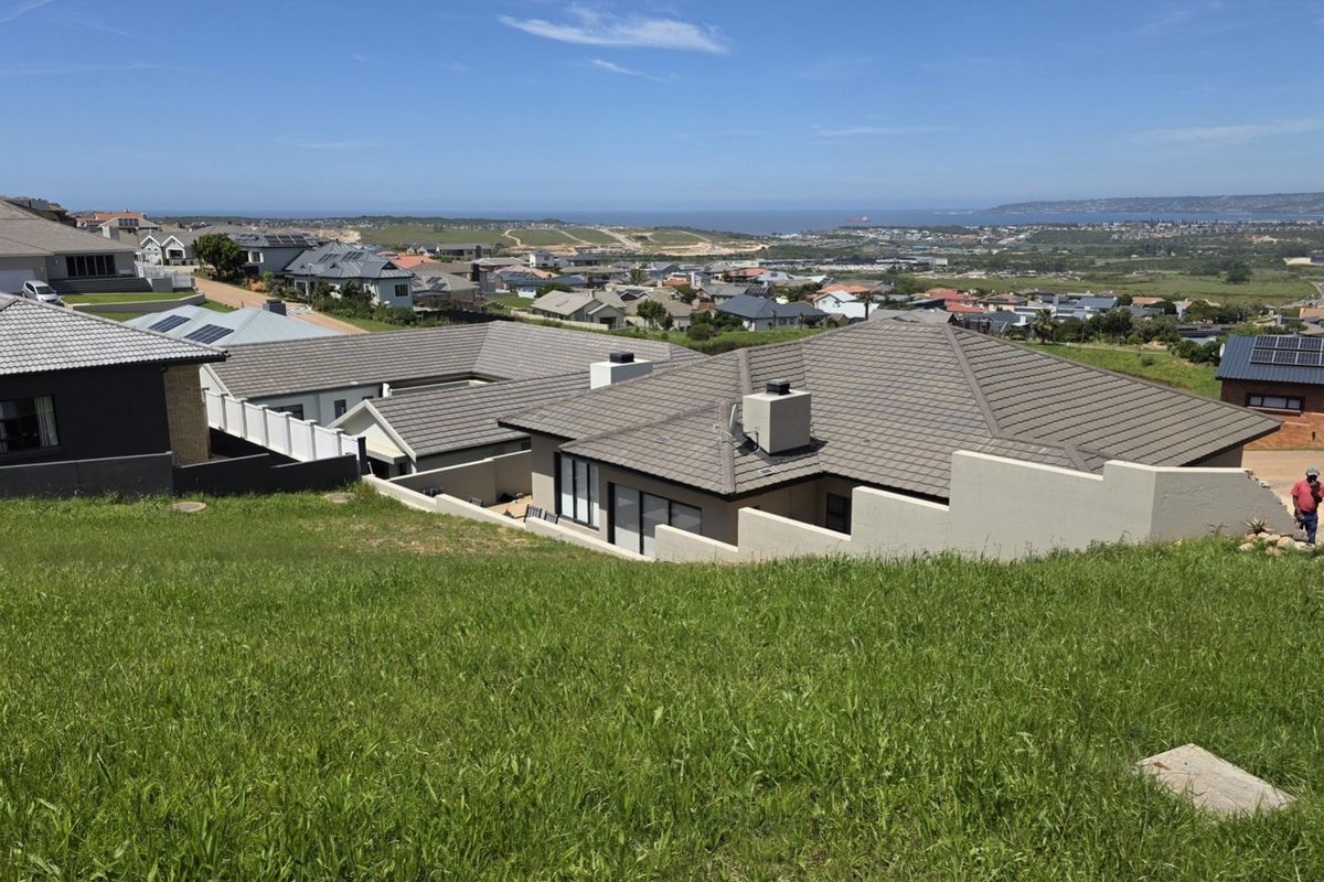 An elevated view of a sloped residential plot overlooking a suburban neighborhood with distant coastal views.