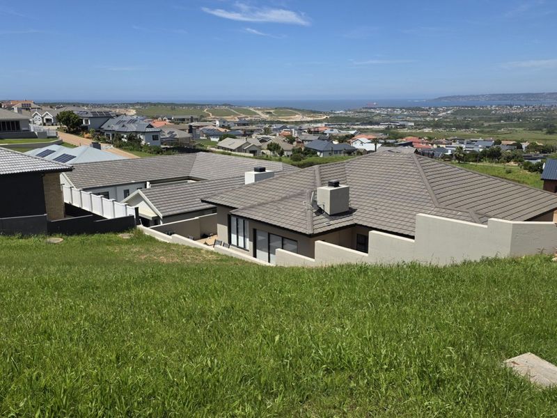 An elevated view of a sloped residential plot overlooking a suburban neighborhood with distant coastal views.