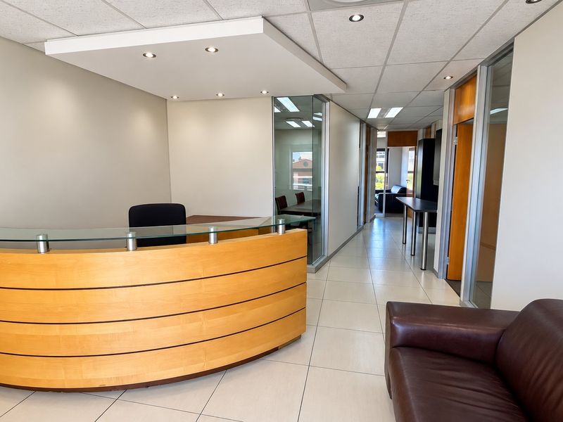 Modern commercial reception area featuring a curved wooden desk, glass office partitions, tiled flooring, and professional recessed ceiling lighting.