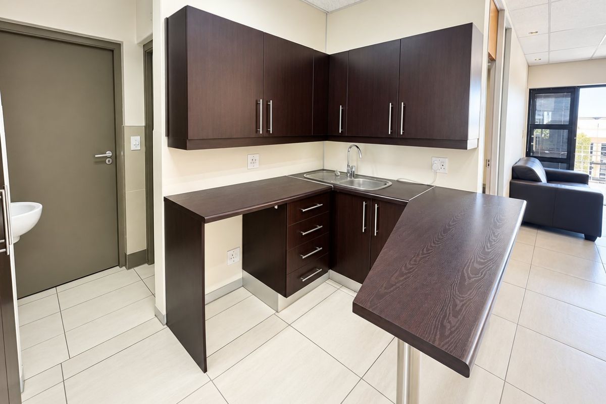 A clean and functional kitchenette featuring dark wood-grain cabinetry, a stainless steel sink, and light-colored tiled flooring in a professional office environment.