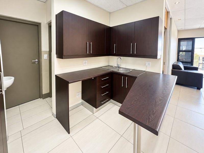 A clean and functional kitchenette featuring dark wood-grain cabinetry, a stainless steel sink, and light-colored tiled flooring in a professional office environment.