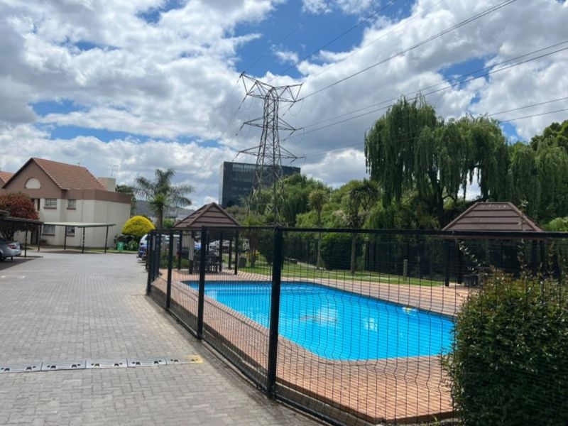 Communal swimming pool area featuring a paved surround, gazebo, and perimeter fencing, situated adjacent to the complex parking and carport facilities.