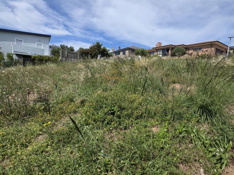 A view of a vacant residential plot featuring a sloped landscape with natural vegetation, situated in an established neighborhood.