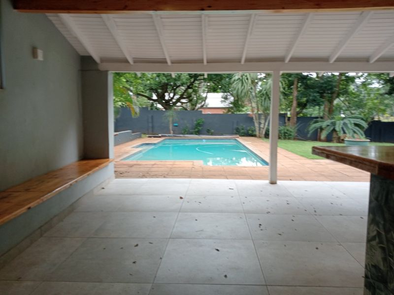 A spacious covered patio featuring a built-in bar and bench, overlooking a private swimming pool and garden area, ideal for outdoor entertaining.