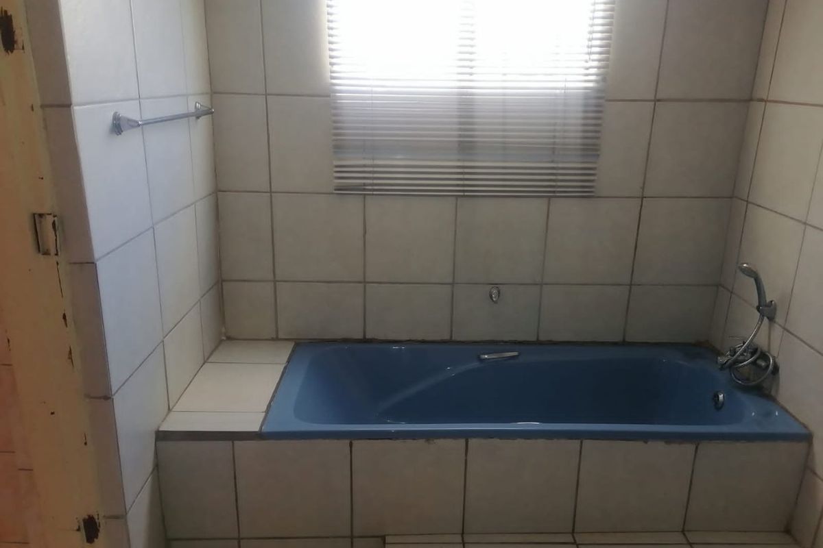 A dated bathroom featuring a built-in blue bathtub, tiled walls and flooring, and a window with fitted blinds.