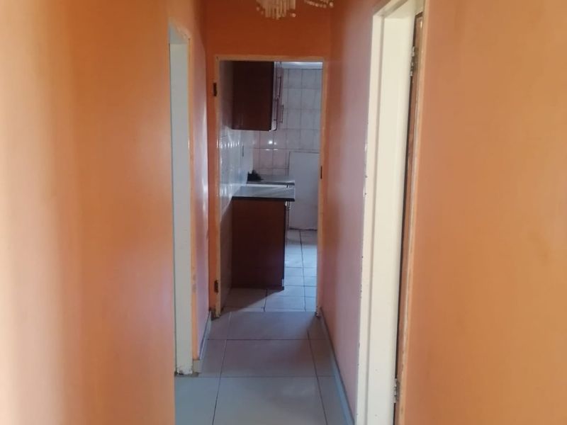 A narrow interior hallway featuring tiled flooring and a decorative chandelier, leading towards a kitchen area with visible cabinetry.