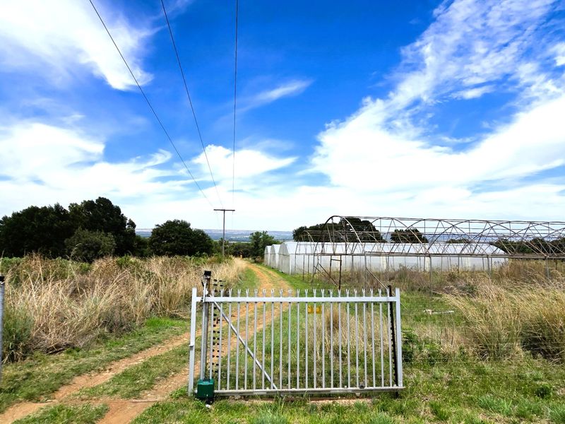 A rural farm property featuring a metal security gate, electric fencing, and multiple greenhouse structures set along a dirt access track.