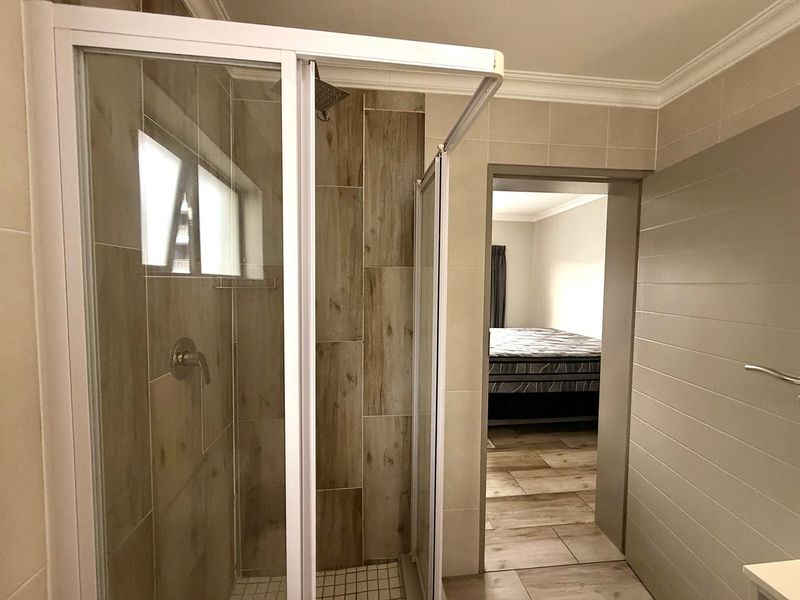 A modern bathroom featuring a glass-enclosed shower with wood-look wall tiles and neutral floor tiling, leading into an adjacent bedroom.