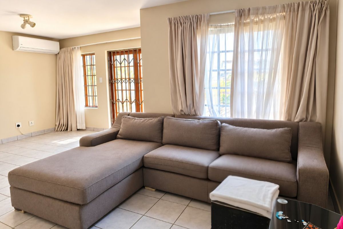 A bright and spacious living room featuring tiled flooring, a wall-mounted air conditioning unit, and secure access points with security gates and window bars.